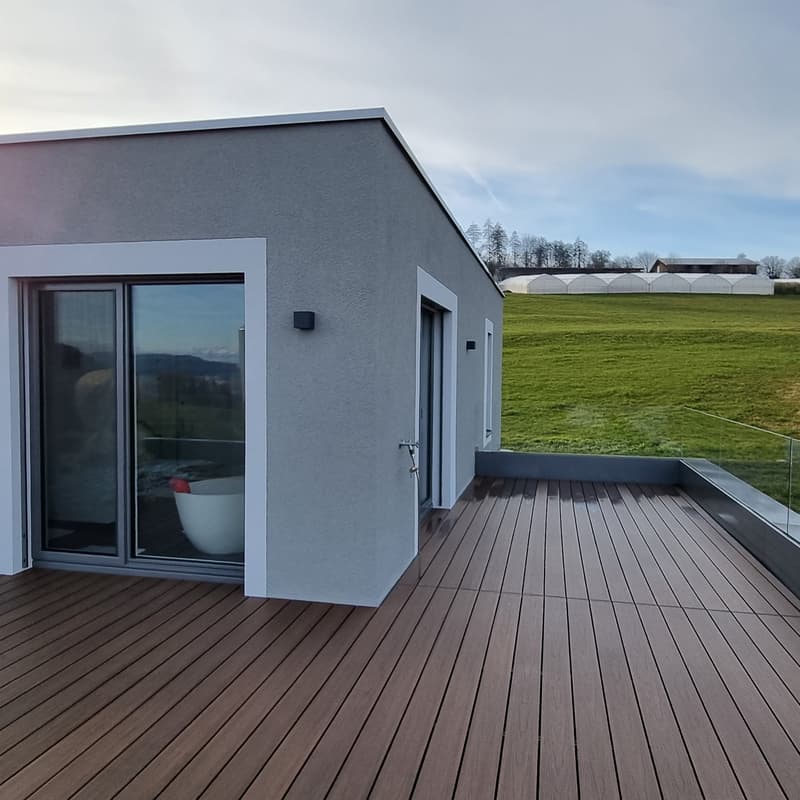 A balcony with a wooden deck, a bathtub inside, glass sliding doors, and an expansive view of a green field.