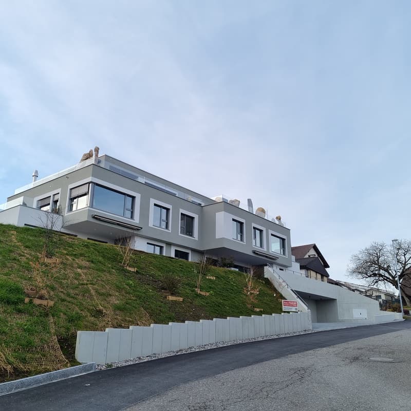 Modern 2 story house, gray walls, large windows, garage entrance, raised ground floor, balcony on the top floor, sloped ground