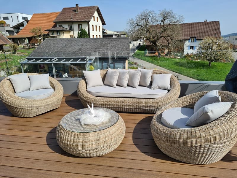 Terrace with wicker furniture, a large glass table, white pillows, and a clear view of the surrounding houses.