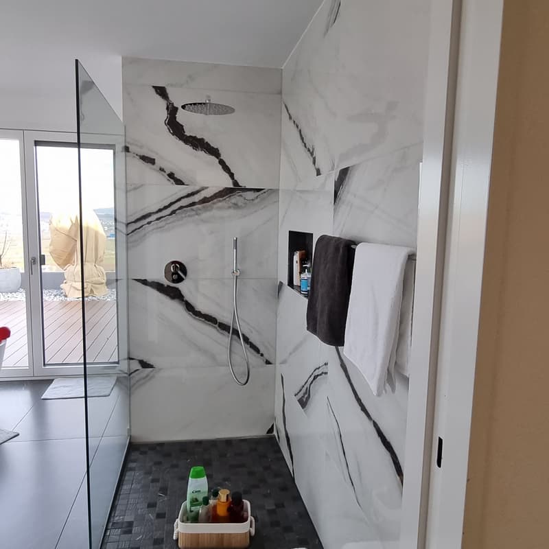 Modern bathroom with white marble walls, black and white tiled floor, glass shower enclosure, glass door with wooden floor, towels, toiletries on a shelf, black and white decor.