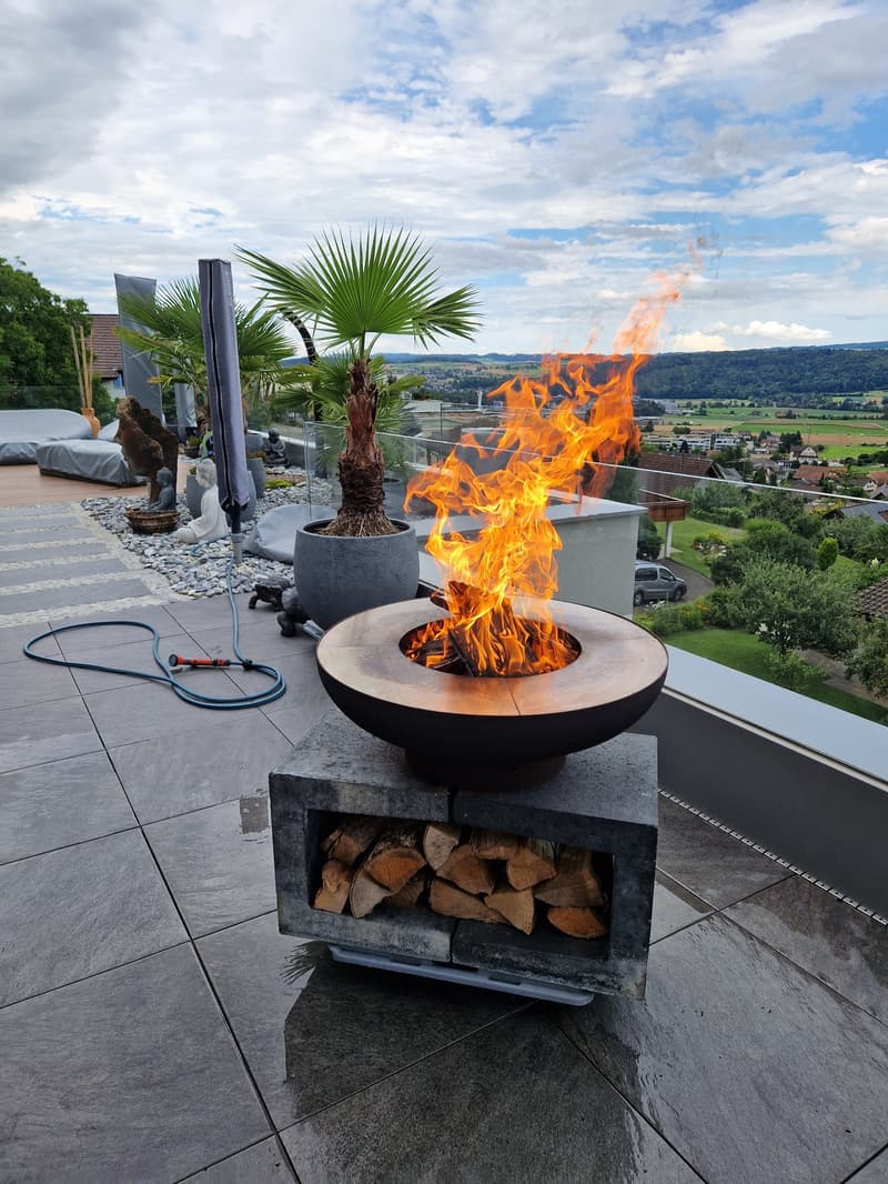 A large fire pit with logs on a concrete base is on a terrace with a fireproof table, palm trees, plants, and a covered area. There is a beautiful view of the surrounding countryside with houses and mountains in the distance.