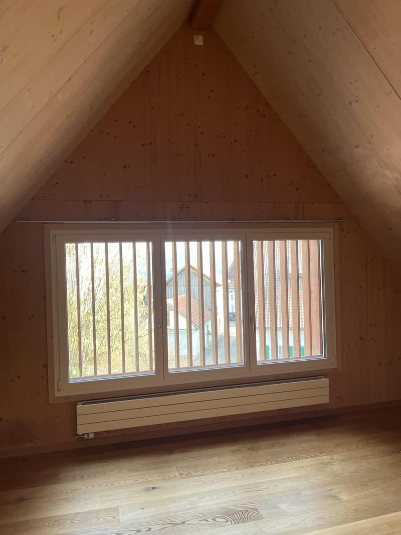 A small attic room with wooden ceiling and walls, a large window and a white radiator