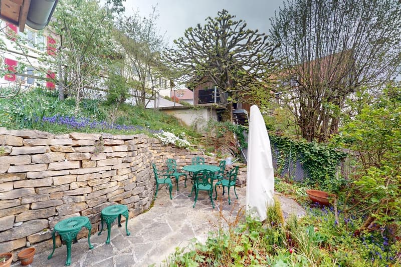 A garden with stone wall, green chairs, table, potted plants, flowers and trees.