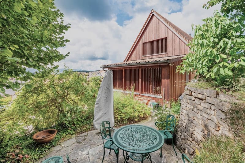 outdoor stone patio, stone wall, green chairs, green table, plants, trees, and a view of a house