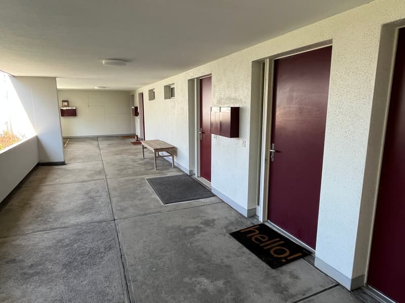 Empty corridor with concrete floor, maroon doors, a bench, and a mat in front of one of the doors.