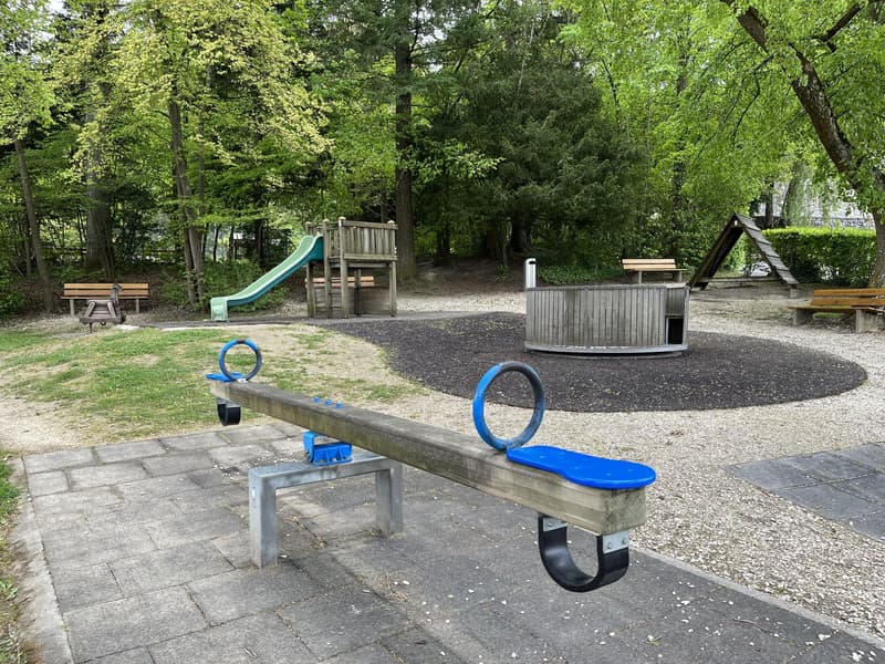 Outdoor playground with a seesaw, slide, and benches, surrounded by trees and grass.