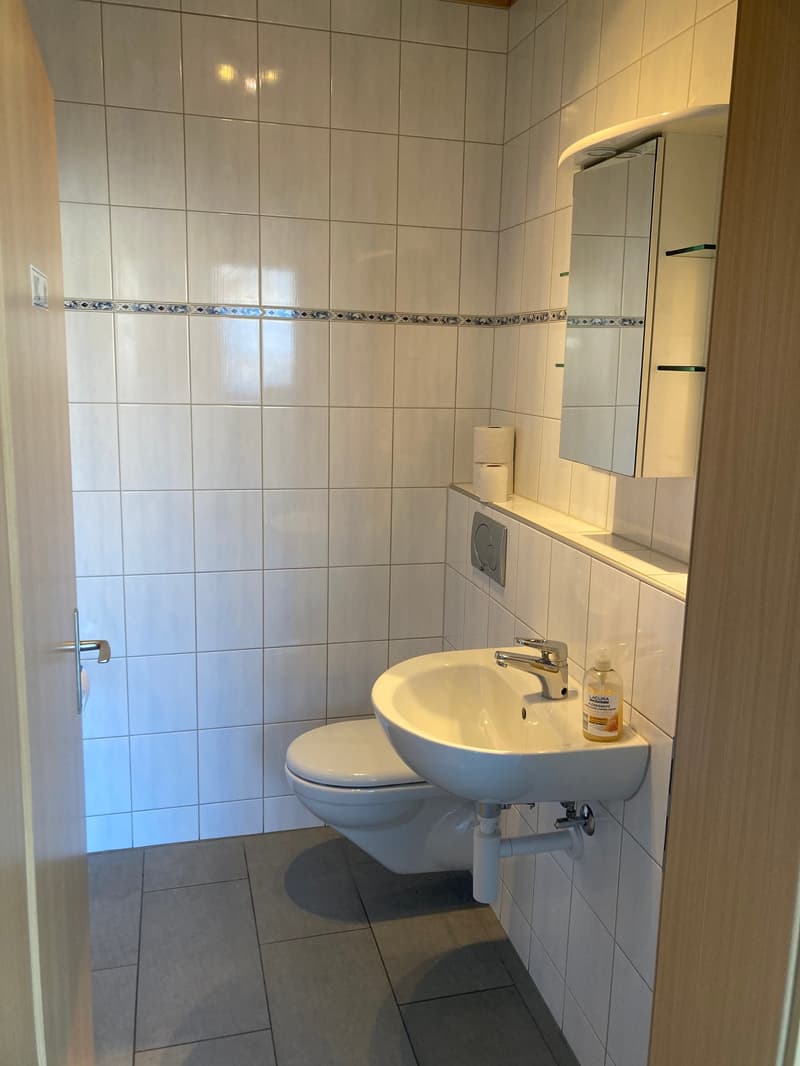 A small bathroom with white tiles, a toilet, sink, and mirror, a wooden door, and a silver faucet