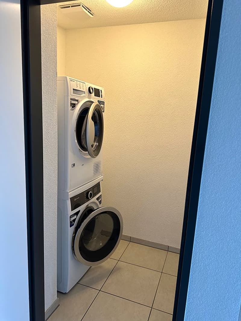 A small room with a stacked washer and dryer, beige walls, and tiled floor.