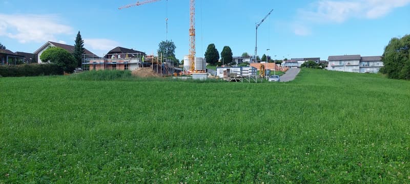 The image shows a construction site with a large crane and several buildings under construction. The site is surrounded by a grassy field and there appears to be a garage or other structure visible in the background.