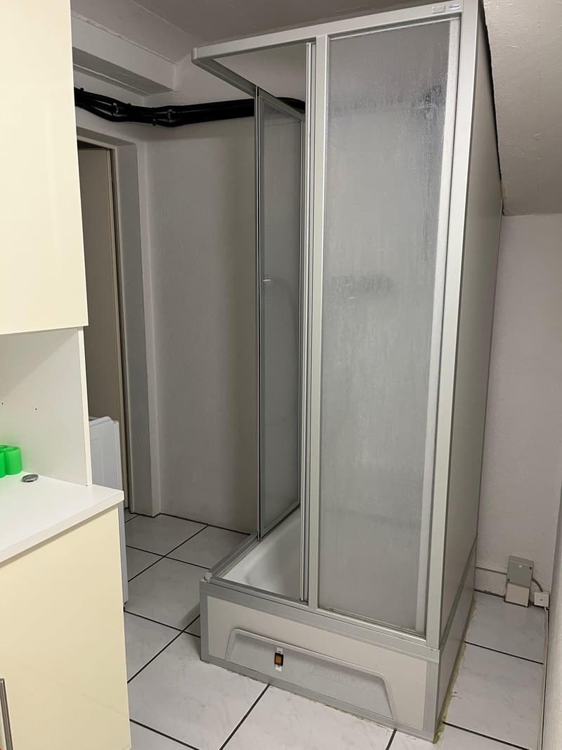 corner shower stall with glass doors, white tile flooring, small shelf with three cups, white walls, a door to another room with washing machine