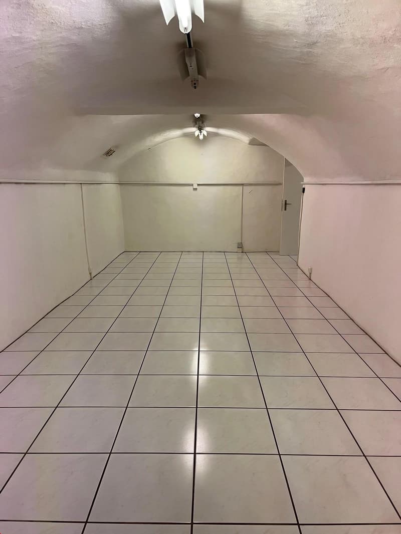 Empty room with white tiled floor, white walls, two entrance doors, ceiling lights, and an open ceiling space with lights