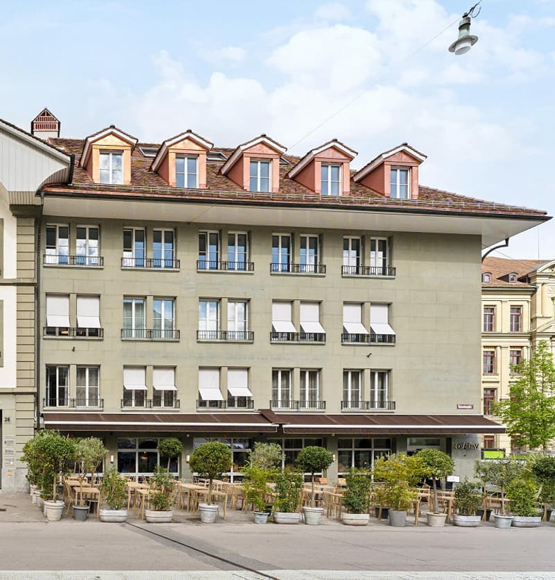 a large building with multiple balconies and windows, ground floor cafe with patio furniture and green trees, outdoor space with lots of potted plants