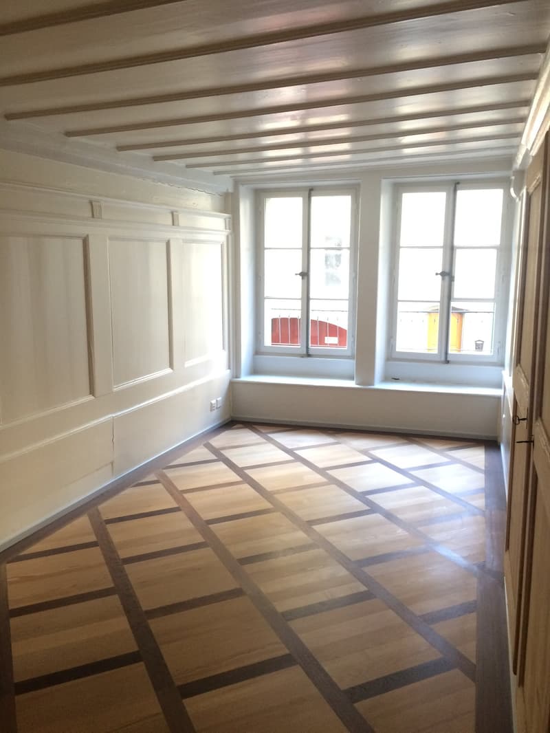 An empty room with hardwood floor, white walls, 2 windows, and window sills.