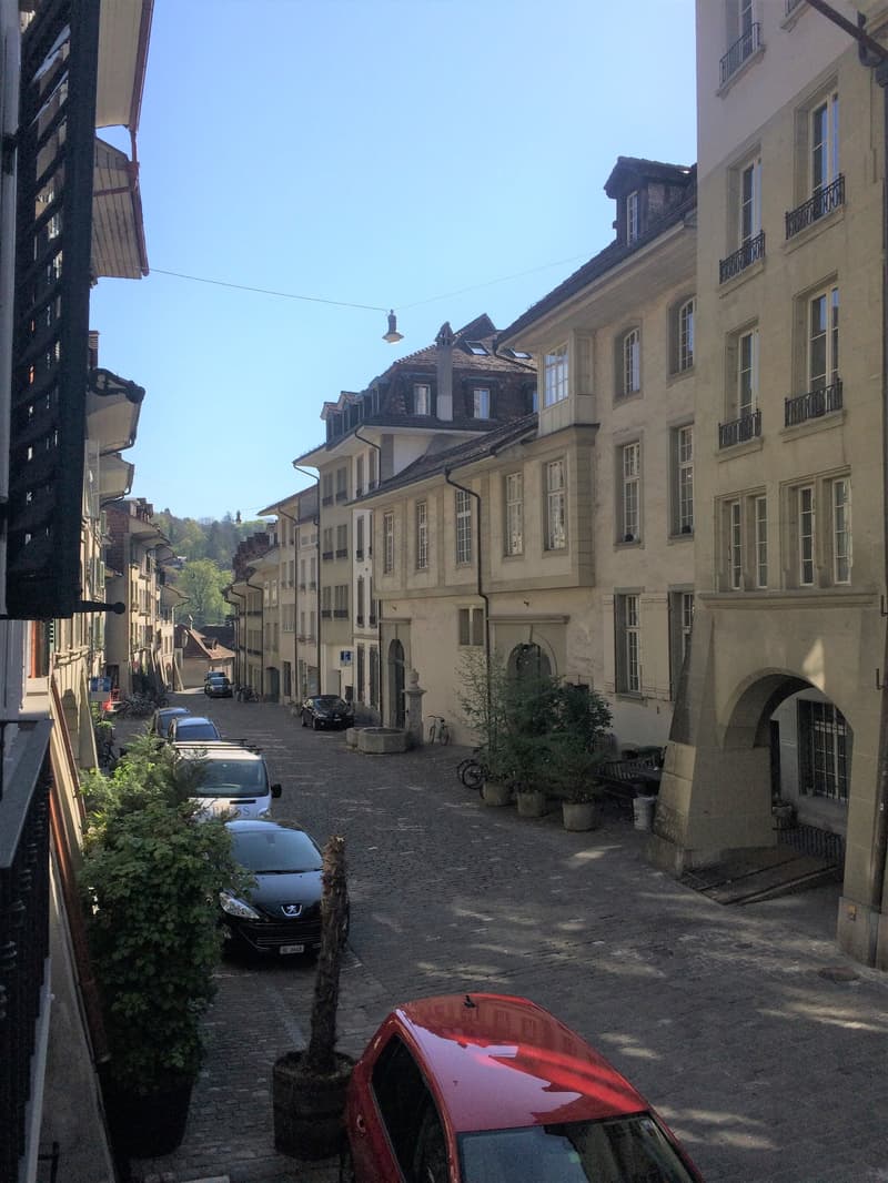 A narrow street with multi-storey buildings on both sides, paved with cobblestones. Some parked cars are visible along the street.