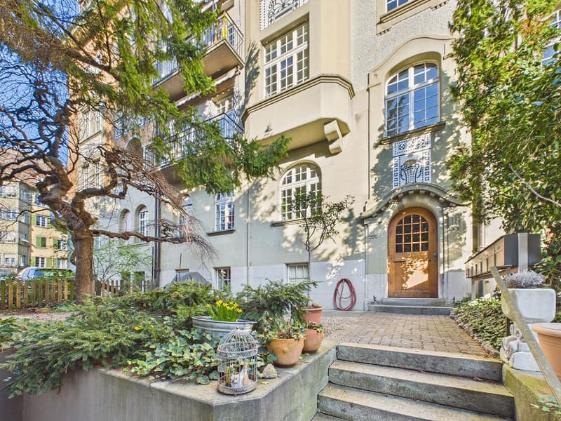 3 story building, beige walls, wooden door, brick pathway, garden with plants and trees