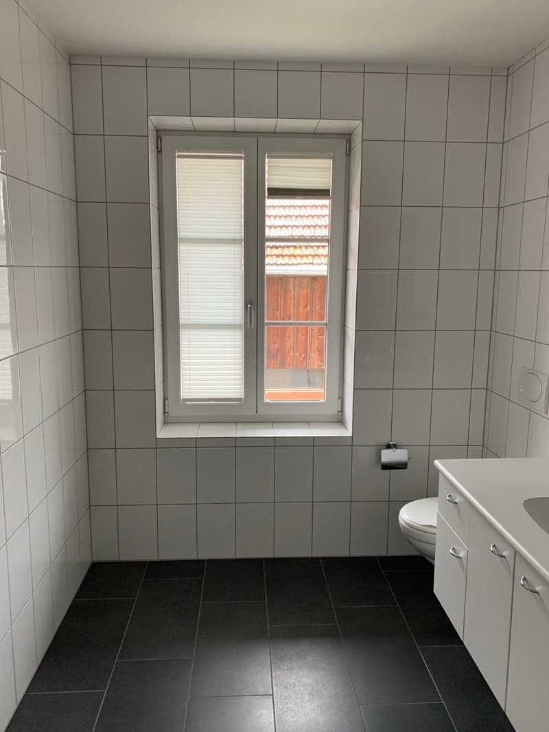 Bathroom with white tiles, black tiles on the floor, window with blinds, toilet, sink, and cabinets