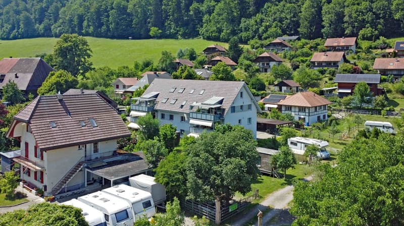 Aerial view of residential houses, a raised ground floor, some with balconies, in a mountainous area with green surroundings.