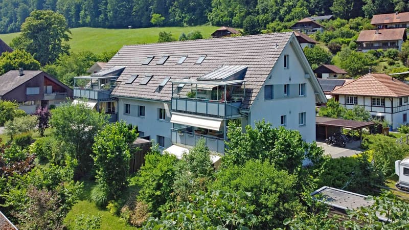 Two-story house with a flat roof and solar panels, surrounded by green trees and bushes. It has multiple balconies and windows.