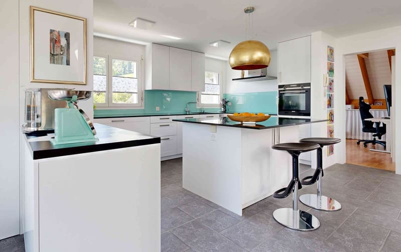 modern kitchen with white cabinets, black countertops, gray floor tiles, center island with bar stools, hanging light fixture, kitchen appliances including an oven and a sink, framed picture on the wall