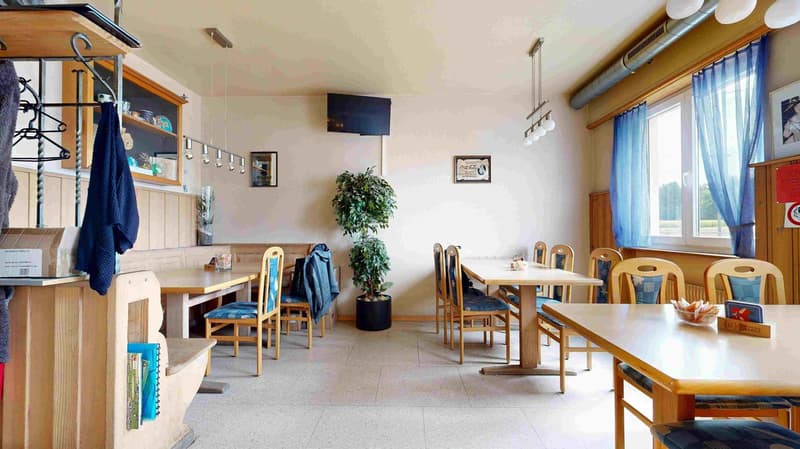 Dining area with wooden tables and chairs, potted plant, TV, blue curtains, wooden shelves, hanging clothes