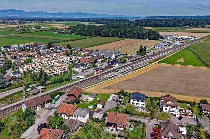 Aerial view of a residential area with houses, a railway track, and surrounding fields and trees.
