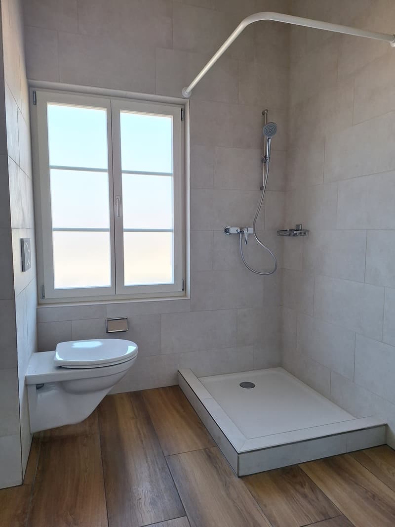 Bathroom with white tiles, wooden floor, white toilet, shower with shower head, window