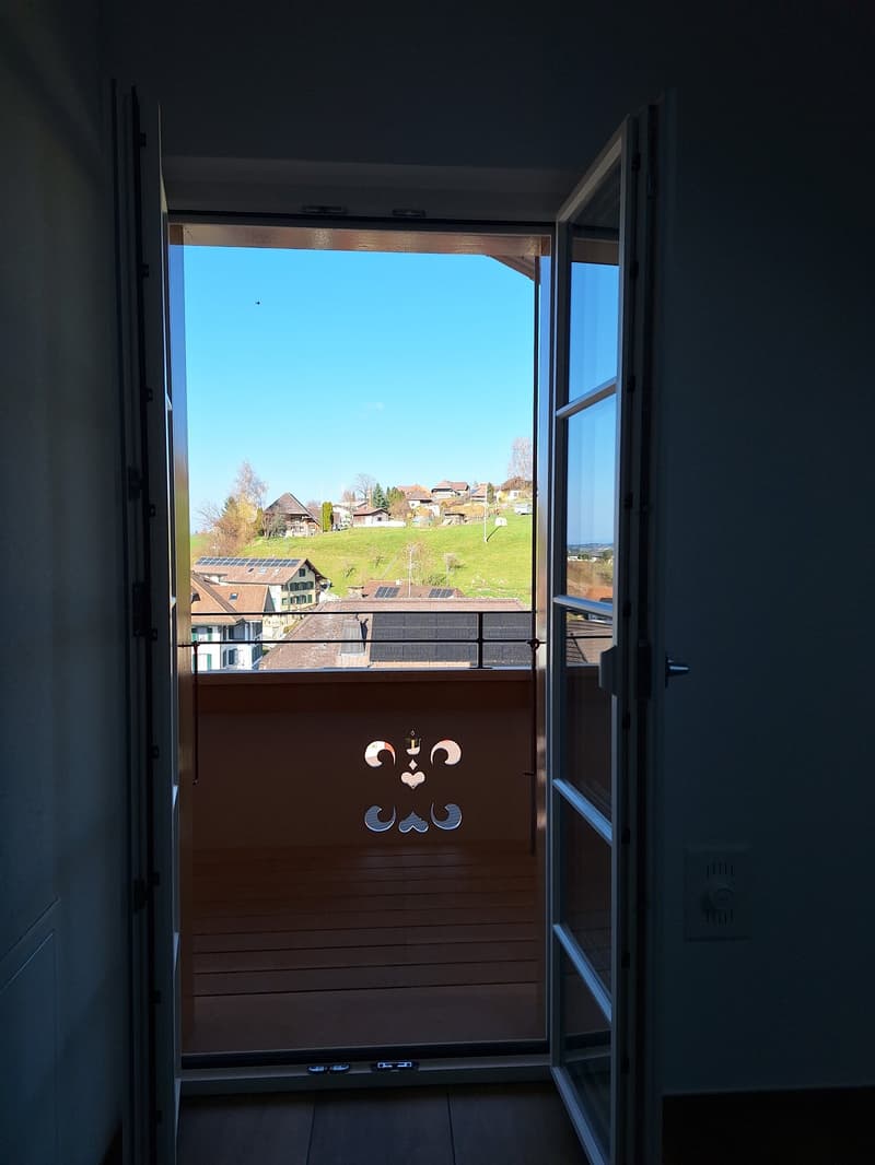 Open door leading to a balcony with a wooden floor and a metal railing, overlooking a green hill with houses and solar panels