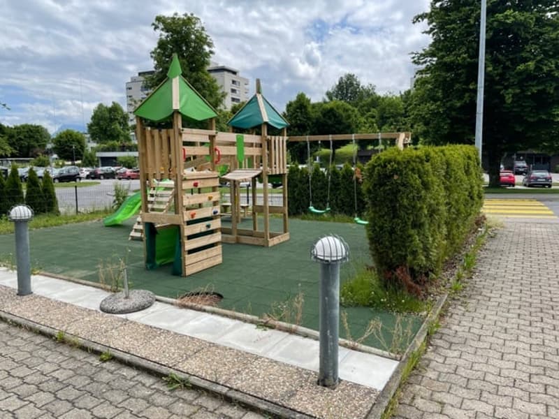 Wooden play structure with slides and swings, green rubber flooring, lamp posts, bushes