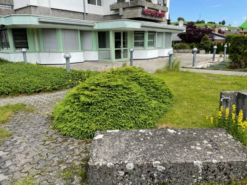The exterior of a 2 story house with concrete and green accents, a garden with bushes and flowers, a concrete pathway, and several poles in front