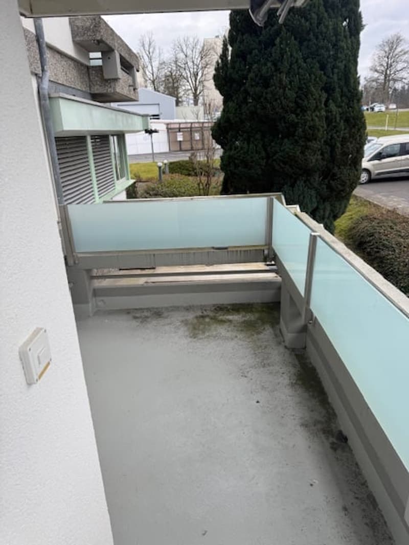 Outdoor balcony with concrete flooring, glass railings, building wall on the left, tree and greenery in the background, vehicles parked in the parking lot, and a car driving on the road.