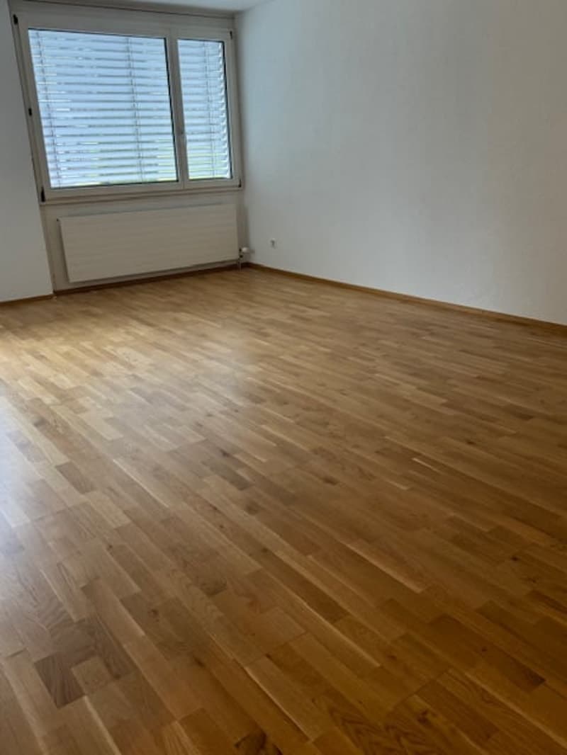 This image shows a spacious room with hardwood flooring, white walls, and a large window with horizontal blinds. The room appears to be empty, providing a clean and minimalist look.