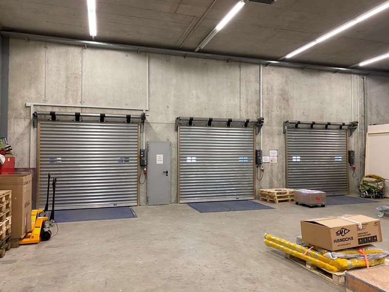 The image shows a warehouse or storage facility with several large rolling garage doors, concrete walls, and overhead lighting. There are pallets and boxes visible, indicating it is used for storage and distribution of goods.