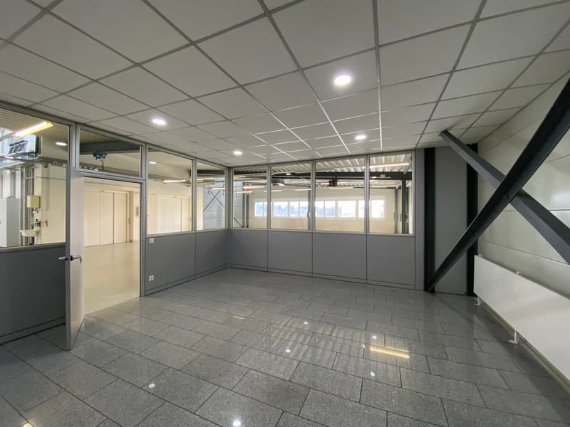 an empty office room with gray flooring, glass walls, metal beams on the ceiling, white ceiling tiles, ceiling lights, an open door leading to another room