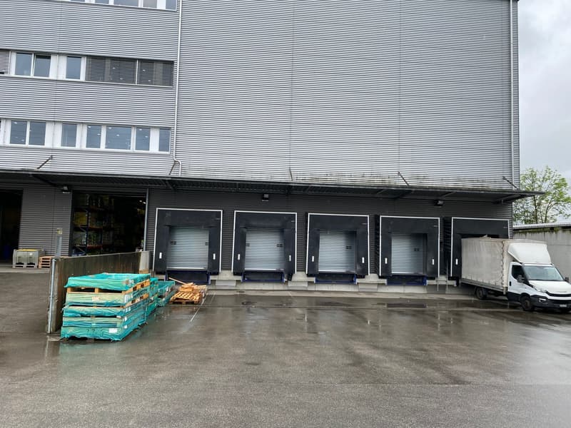The image shows a large industrial building with multiple loading dock doors. There are stacks of pallets and a delivery van parked outside, indicating this is likely a warehouse or distribution center.