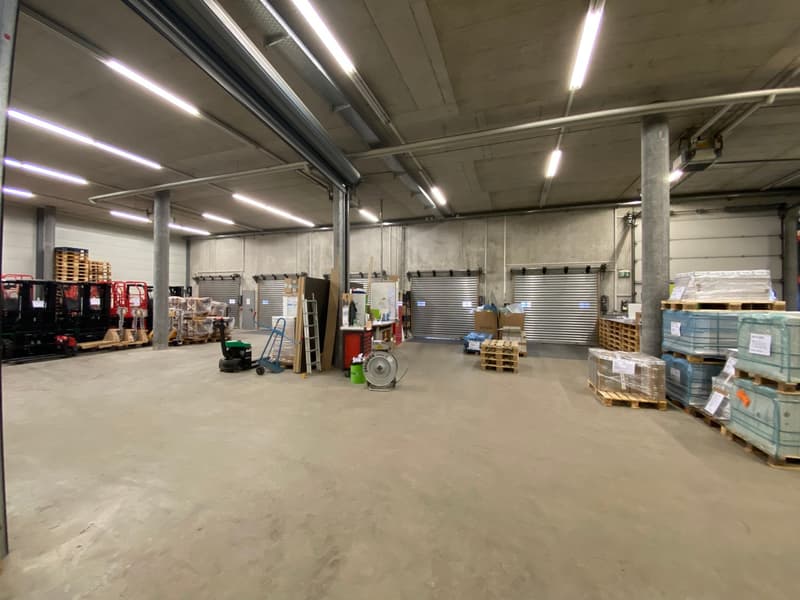 Large industrial warehouse space with concrete floors, metal shelving units, pallets, and various equipment and supplies stored throughout the space.