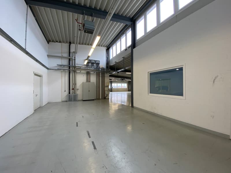 An industrial space with white walls and a high ceiling, illuminated by large windows and ceiling lights, featuring an open area, a door, and several electrical cabinets. The floor appears to be concrete with no furnishings visible.