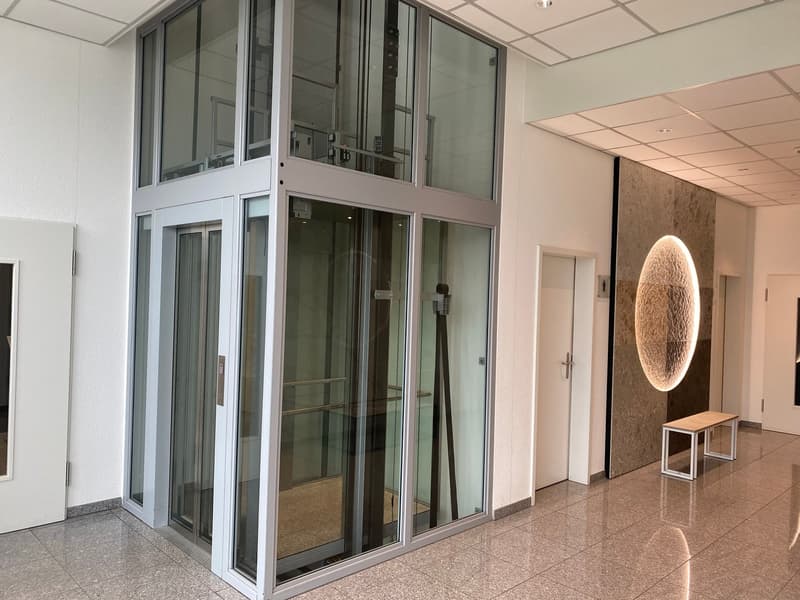 The image shows an interior office space with a glass elevator or lift in the center. The walls are white, and the floor is made of granite or a similar material. There is a large circular light fixture on the wall, and some small benches or tables near t
