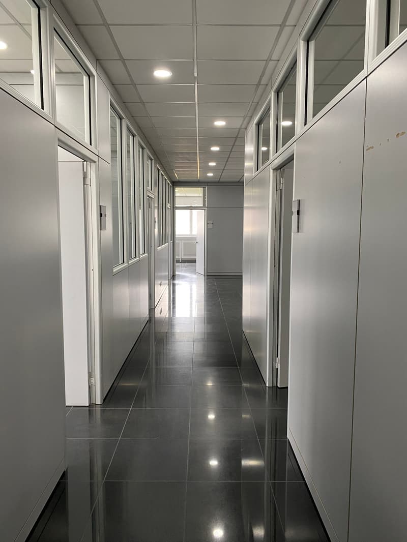 Empty hallway with gray tile flooring, white walls, glass windows, and built-in lights.