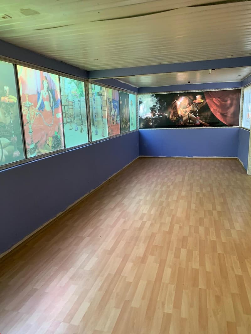 This image shows a large room with a wooden floor and blue walls. The room has several large windows along the walls, displaying various colorful paintings or artwork. There is also a large projection screen or display on one of the walls, showing what ap