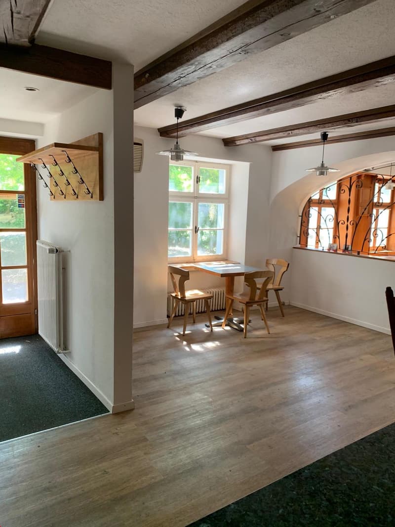 Rustic living room with exposed wooden beams on the ceiling, hardwood floors, a fireplace, and a dining table with wooden chairs. The room has large windows that let in natural light and a cozy, inviting atmosphere.