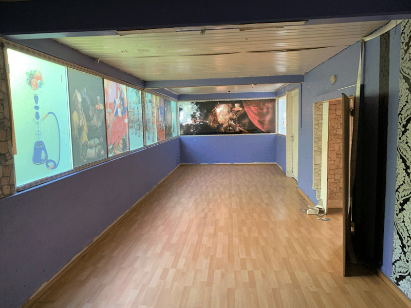 This image shows a large room with a wooden floor and painted walls. The room has a curved ceiling with wooden beams, and there are several framed artworks or paintings displayed on the walls. The room appears to be a multipurpose space, possibly used for