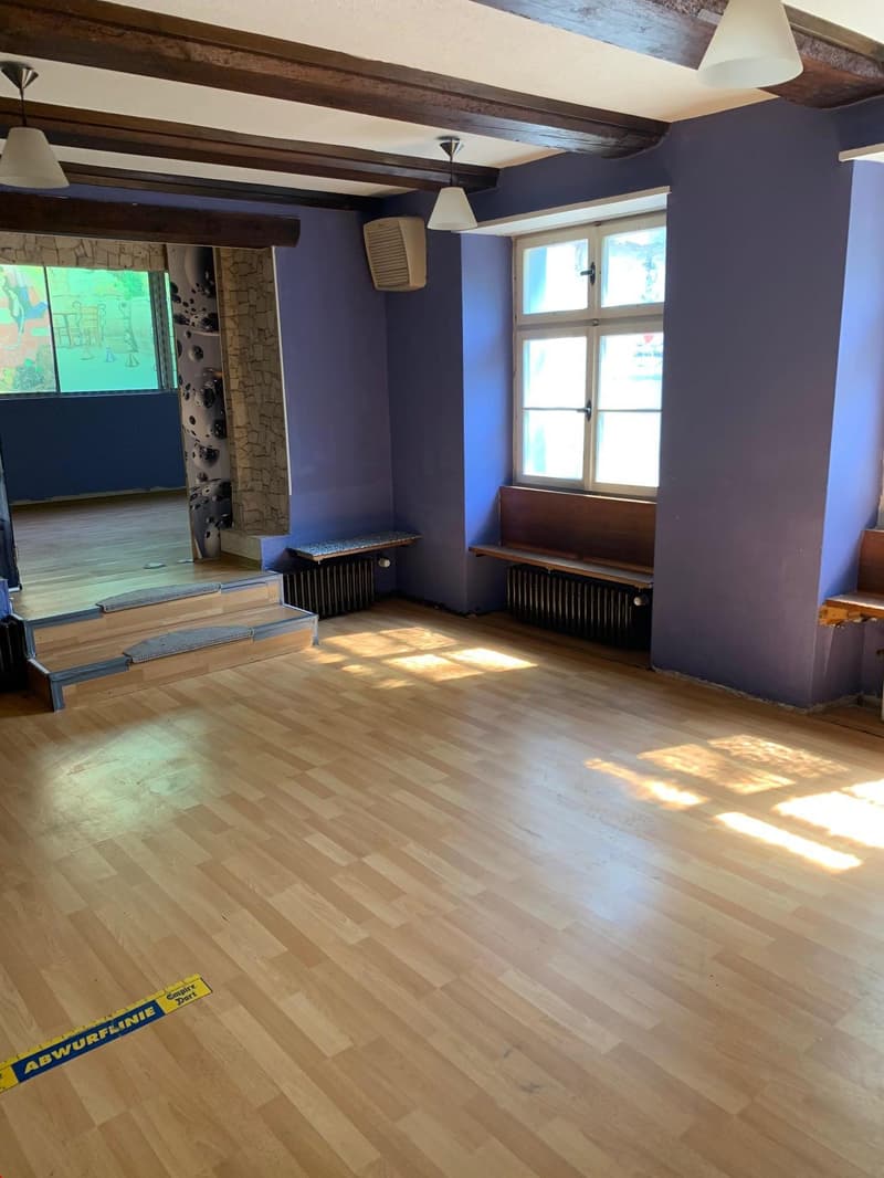Large living room with hardwood floors, exposed wooden beams on the ceiling, and a fireplace. The room has large windows that let in natural light. There are benches along the walls and the space appears to be set up for some kind of activity or event.