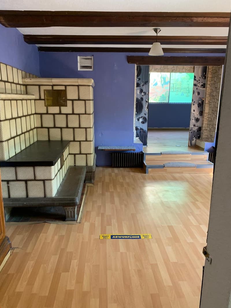 The image shows a living room with a brick fireplace, hardwood floors, and exposed wooden beams on the ceiling. The room has a blue wall color and a window overlooking an outdoor area.
