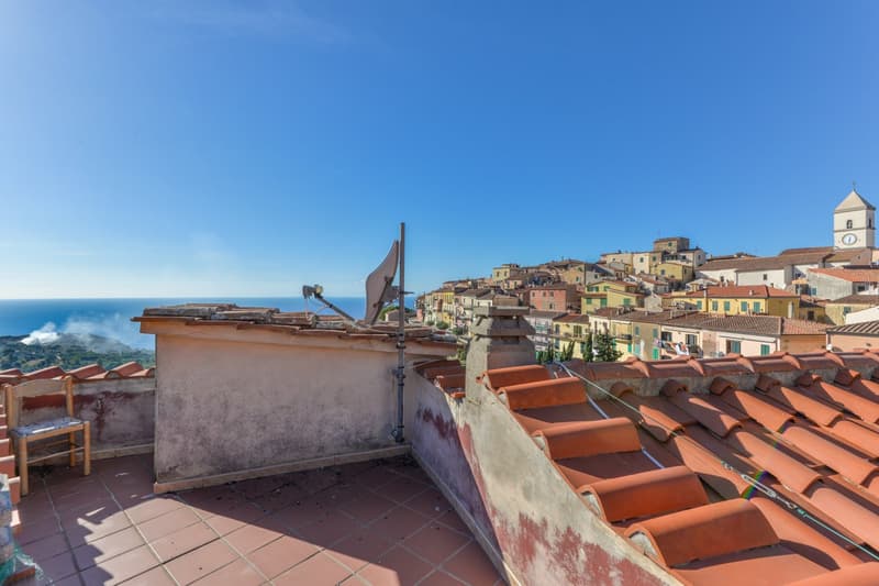 The image shows a rooftop view of a colorful coastal town with traditional Mediterranean-style buildings and red tile roofs. The rooftops are visible in the foreground, and the town is situated on a hillside overlooking the ocean in the distance.
