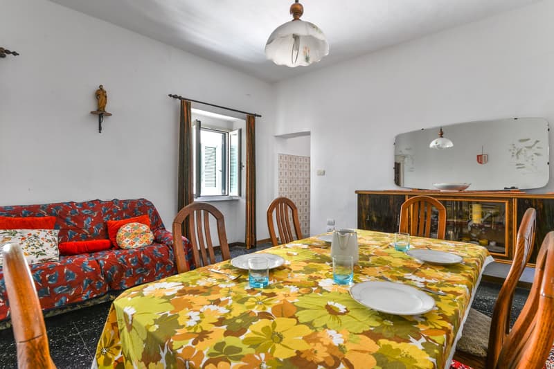 The image shows a combined living room and dining area. The living room has a colorful floral patterned sofa, and the dining area has a large floral tablecloth on the table. There is a wooden cabinet or sideboard in the room, and a decorative wall sconce.