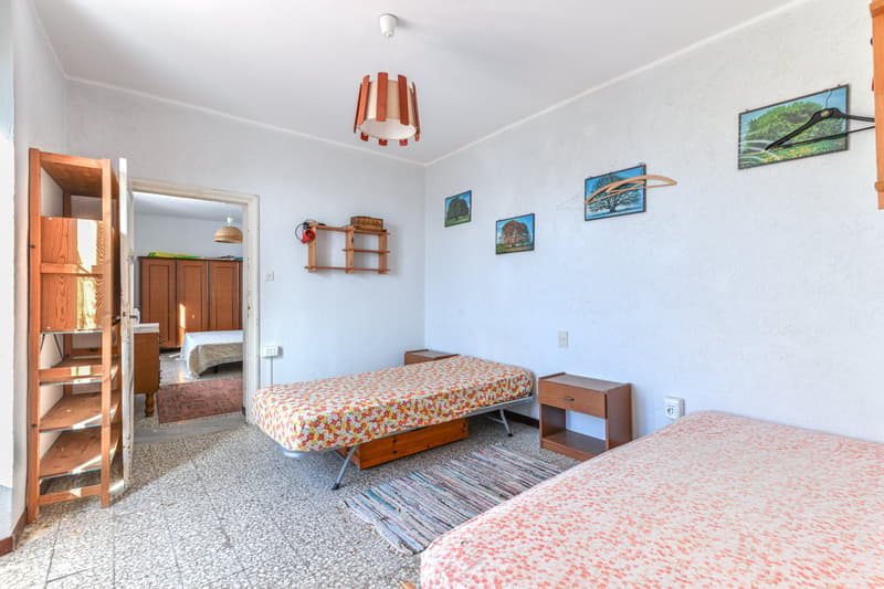 The image shows a bedroom with two twin beds, a wooden shelving unit, and various decorative elements such as framed artwork on the walls. The room has a cozy and casual feel, with a patterned bedspread and a rug on the floor.