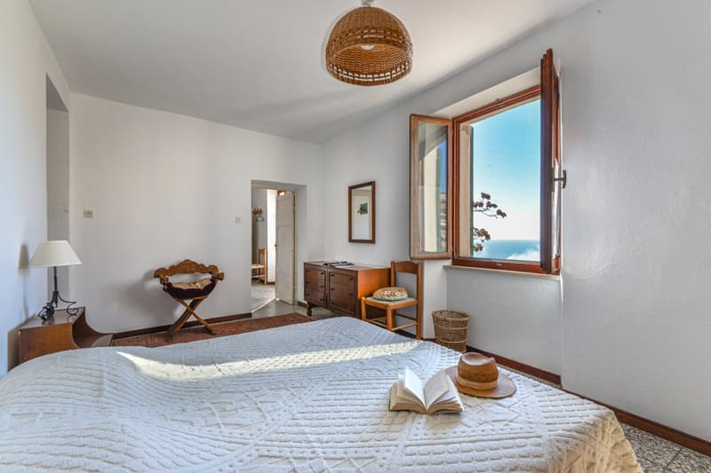 Spacious bedroom with large window offering a scenic ocean view, wicker pendant light, wooden furniture including a dresser and chair, and a cozy bed with a patterned quilt