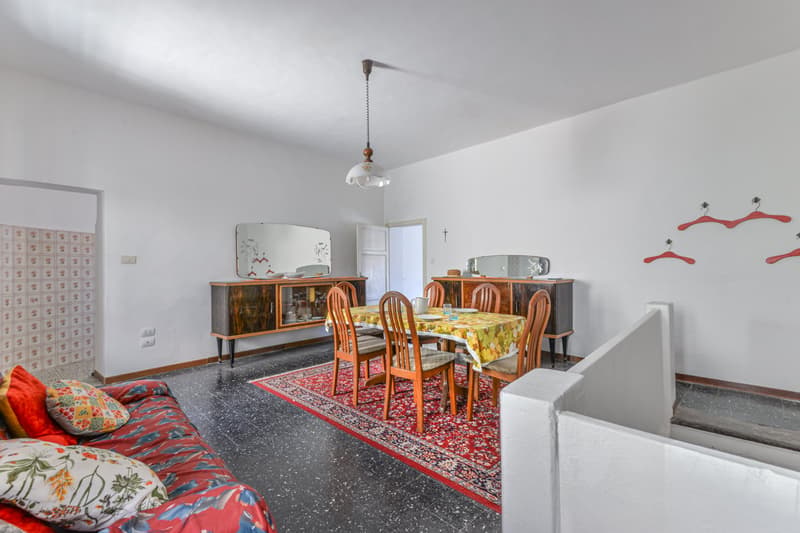 The image shows a combined living room and dining room area. The room has white walls and a tiled floor. There is a large wooden cabinet or sideboard along one wall, with a mirror above it. The dining table is in the center of the room, with wooden chairs