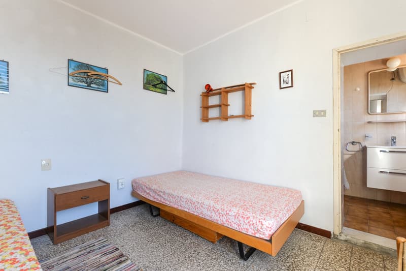 Small bedroom with a single bed, wooden nightstand, and shelving unit. The room has white walls and a tiled floor. There are framed nature photos on the walls.