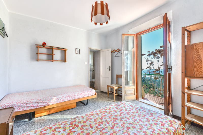 Bedroom with wooden furniture, including a bed frame and shelves. The room has a balcony with a view of the outdoors. The floor is covered with a patterned rug.